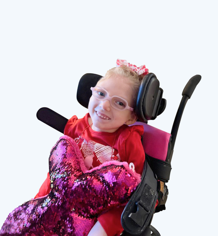 Claire sits in her wheelchair, she has her hair in a scrunchie, is wearing pink glasses and smiling at the camera with a sparkly pink mermaid tail in her lap.