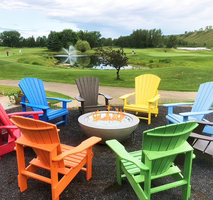 7 colourful wooden lawn chairs surrounding a concrete fire bowl outside.